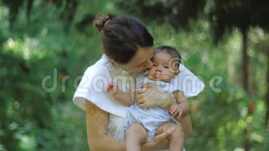 母亲亲吻她的宝贝，手牵着孩子在绿色的草地和树木背景上。 年轻的母亲抱着她